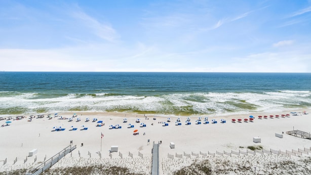 A beach with rows of umbrellas and beach chairs, a wooden walkway, and a fence along the shoreline.