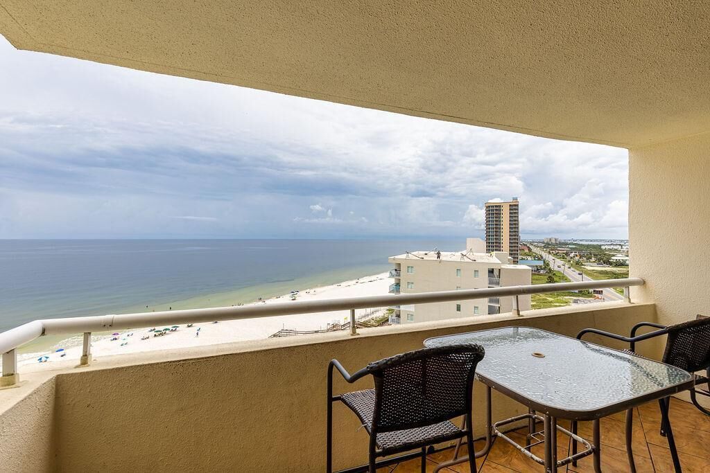 A balcony with a glass table and chairs, overlooking a beach and a tall building.
