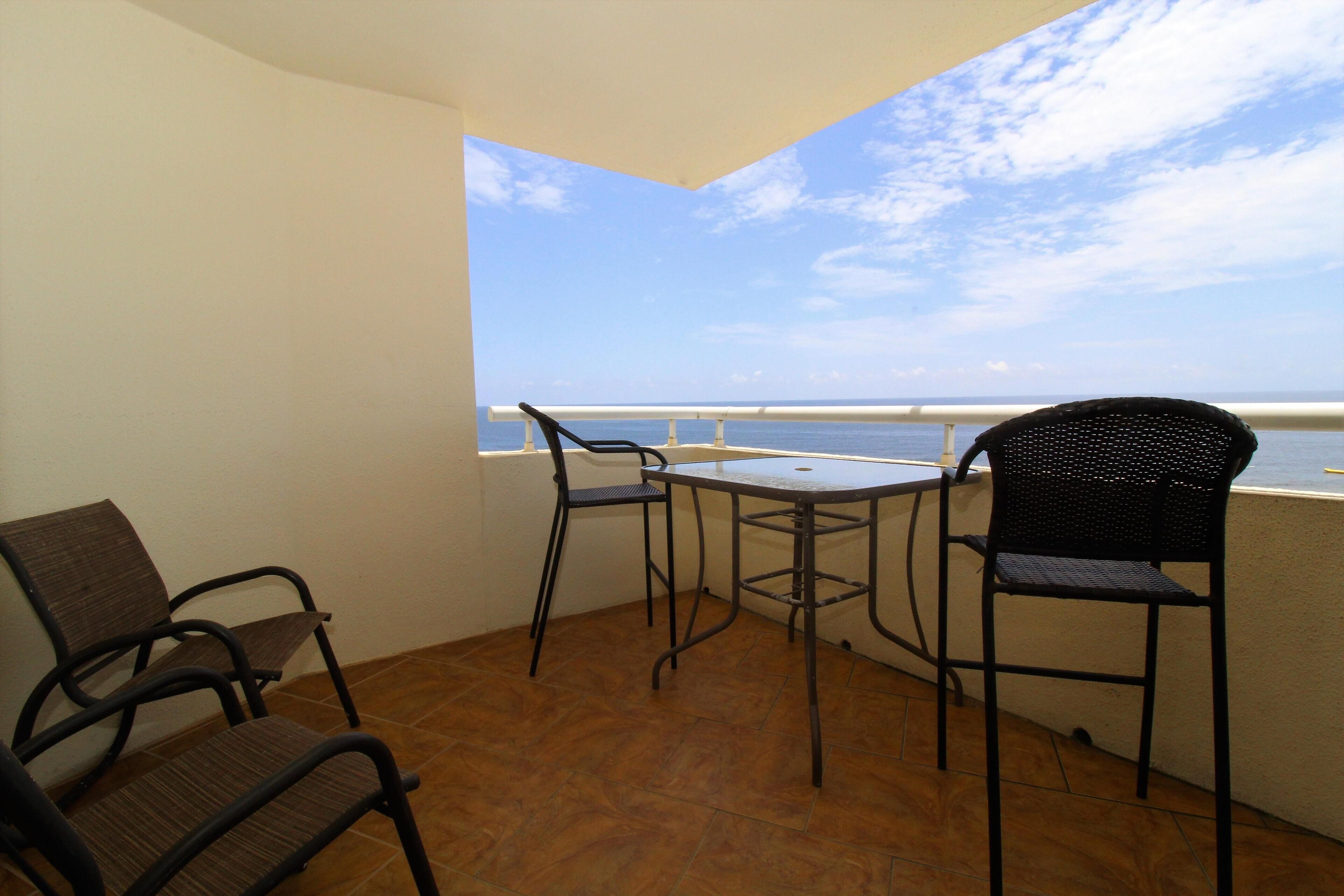 A balcony with a table and chairs, a railing, and a view of the sea.