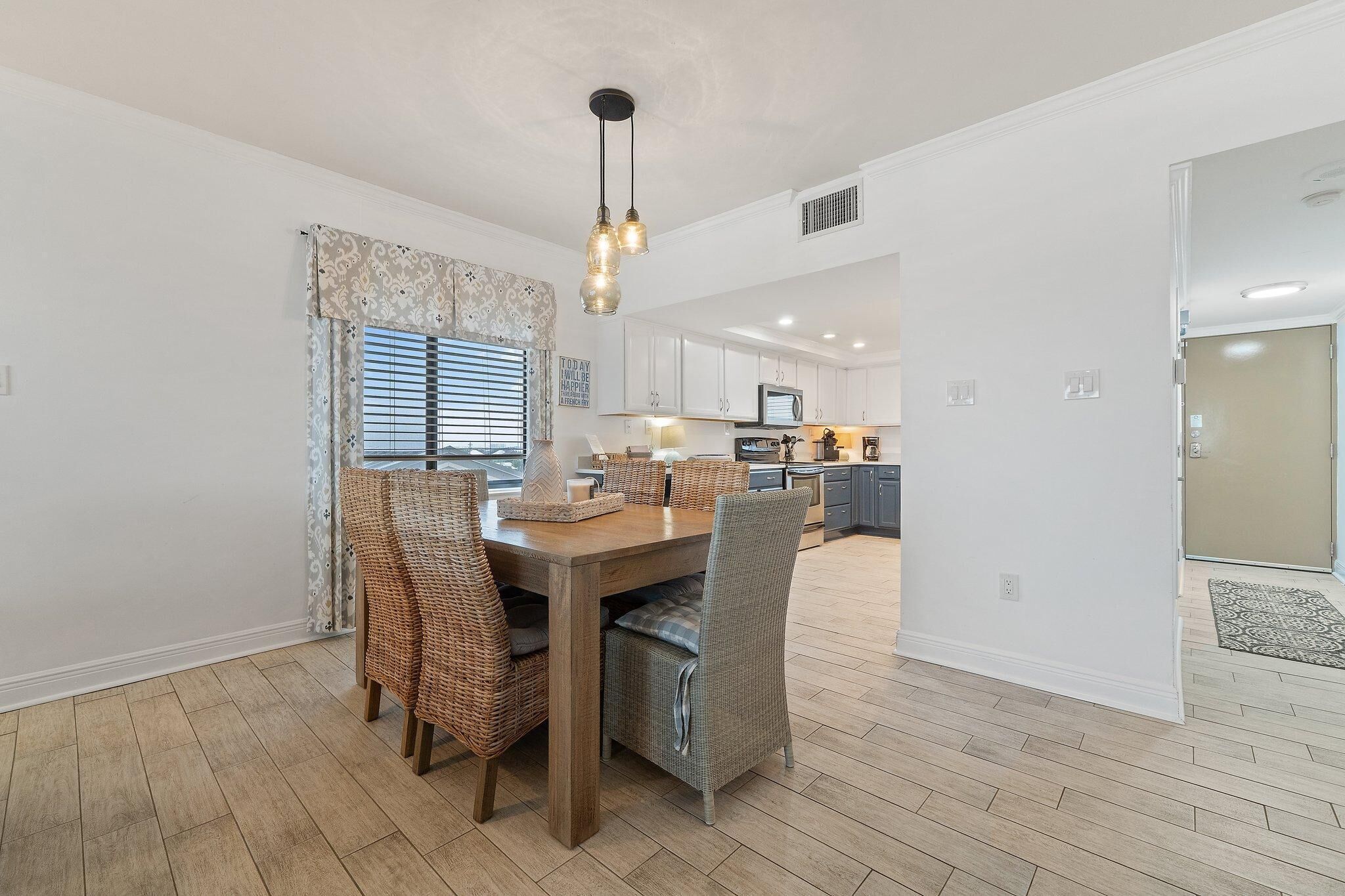 A spacious dining area with a wooden table and wicker chairs, a kitchen in the background, and a view of the outdoors through large windows.