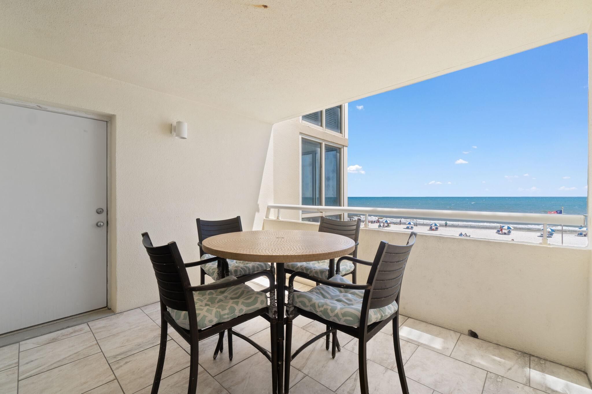 A balcony with a round table and chairs, a door, and a view of the beach and ocean.