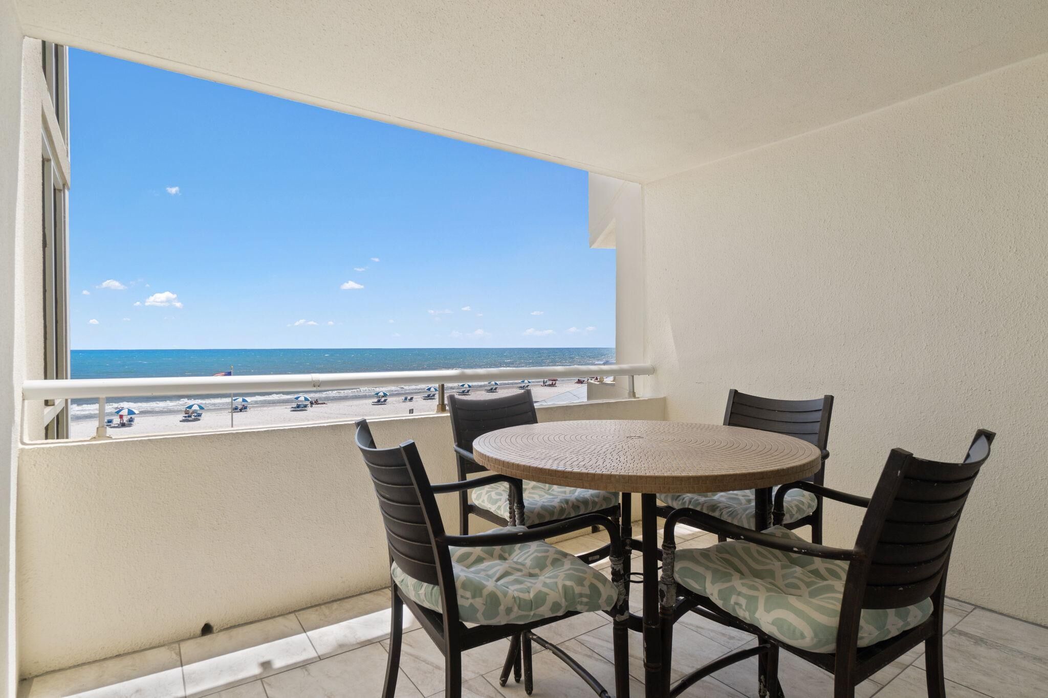 A balcony with a round table and chairs, overlooking a beach with umbrellas and people.