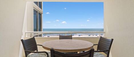 A balcony with a round table and four chairs, overlooking a beach with people and umbrellas.