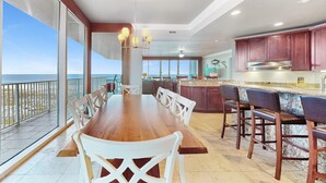 Un vaste espace à manger avec une grande table en bois et des chaises blanches, une cuisine dotée de meubles en bois foncé et une vue sur l’océan grâce à de grandes fenêtres.