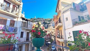 Una calle estrecha flanqueada por edificios, con un balcón adornado con una planta en maceta, y un cielo azul despejado.