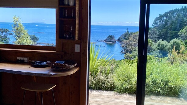 A wooden interior with a large sliding glass door leading to a deck overlooking a coastal area with rocky outcrops and greenery.