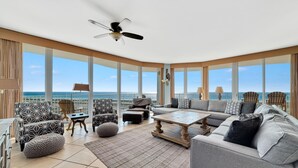 A spacious living area with a large window overlooking the sea, a ceiling fan, and a variety of seating options including a sofa, chairs, and ottomans. - Caribe D1110 (Orange Beach)