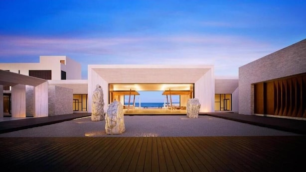 Un hotel moderno con un amplio espacio exterior que cuenta con una terraza de madera, un área de descanso central con vista al mar y un diseño arquitectónico minimalista.
