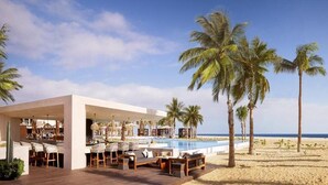 Zona de bar moderna al aire libre con sillones de descanso y piscina, rodeada de palmeras y vista a la playa.