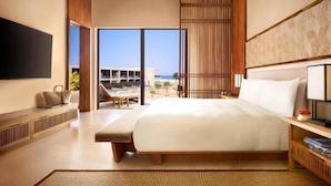 Una habitación de hotel moderna con una cama grande, un televisor de pantalla plana, un área de descanso con vistas a la playa y un ventanal amplio.