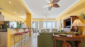 A spacious living area with a green sofa, a wooden dining table, and a kitchen area with a refrigerator and bar stools. - La Playa 502 (Pensacola)