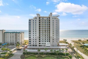 A high-rise hotel with multiple balconies, a parking area, and a view of the beach.