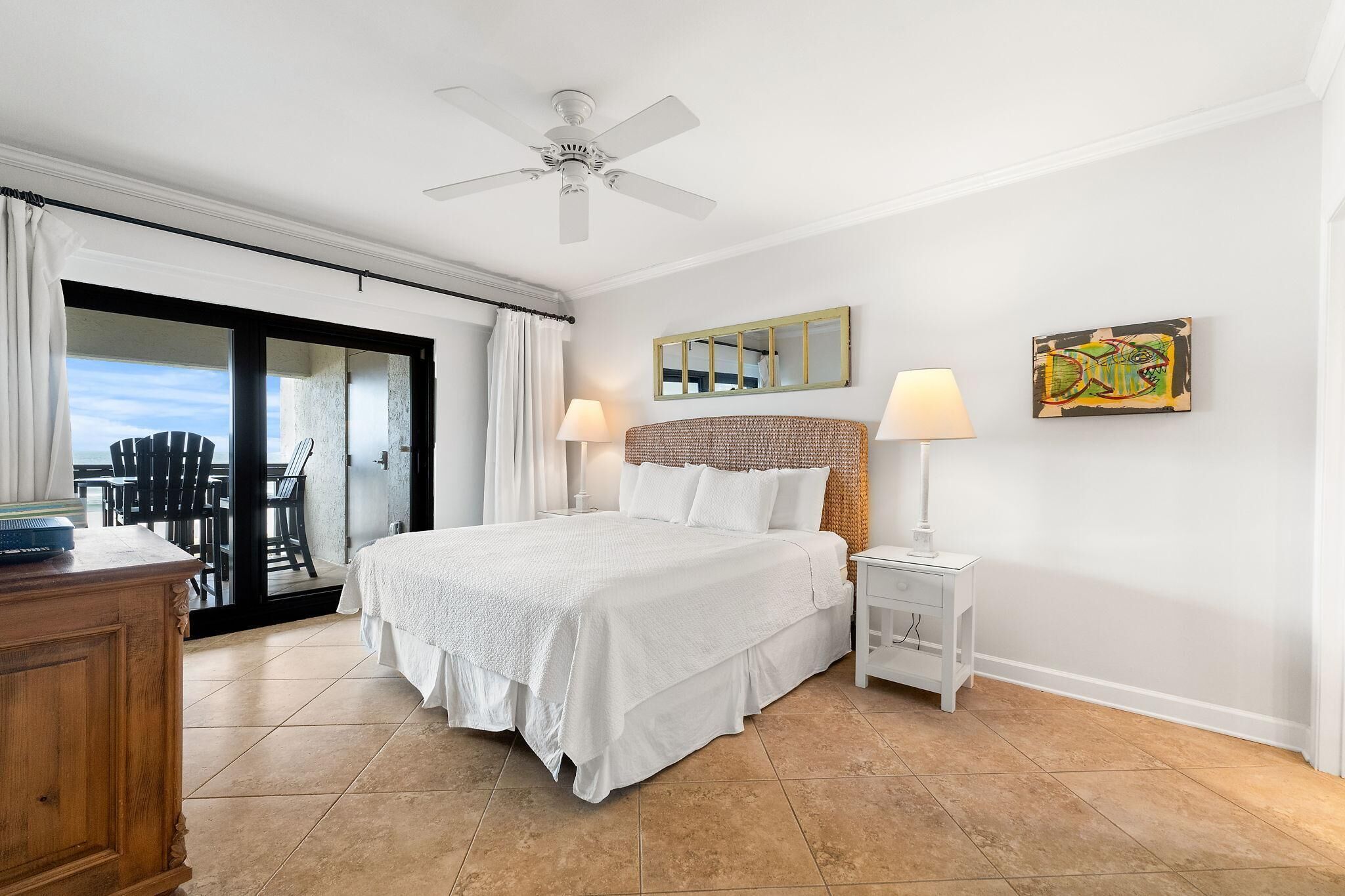 A bedroom with a bed, a nightstand, a lamp, a ceiling fan, a large window with curtains, and a view of the outdoors.