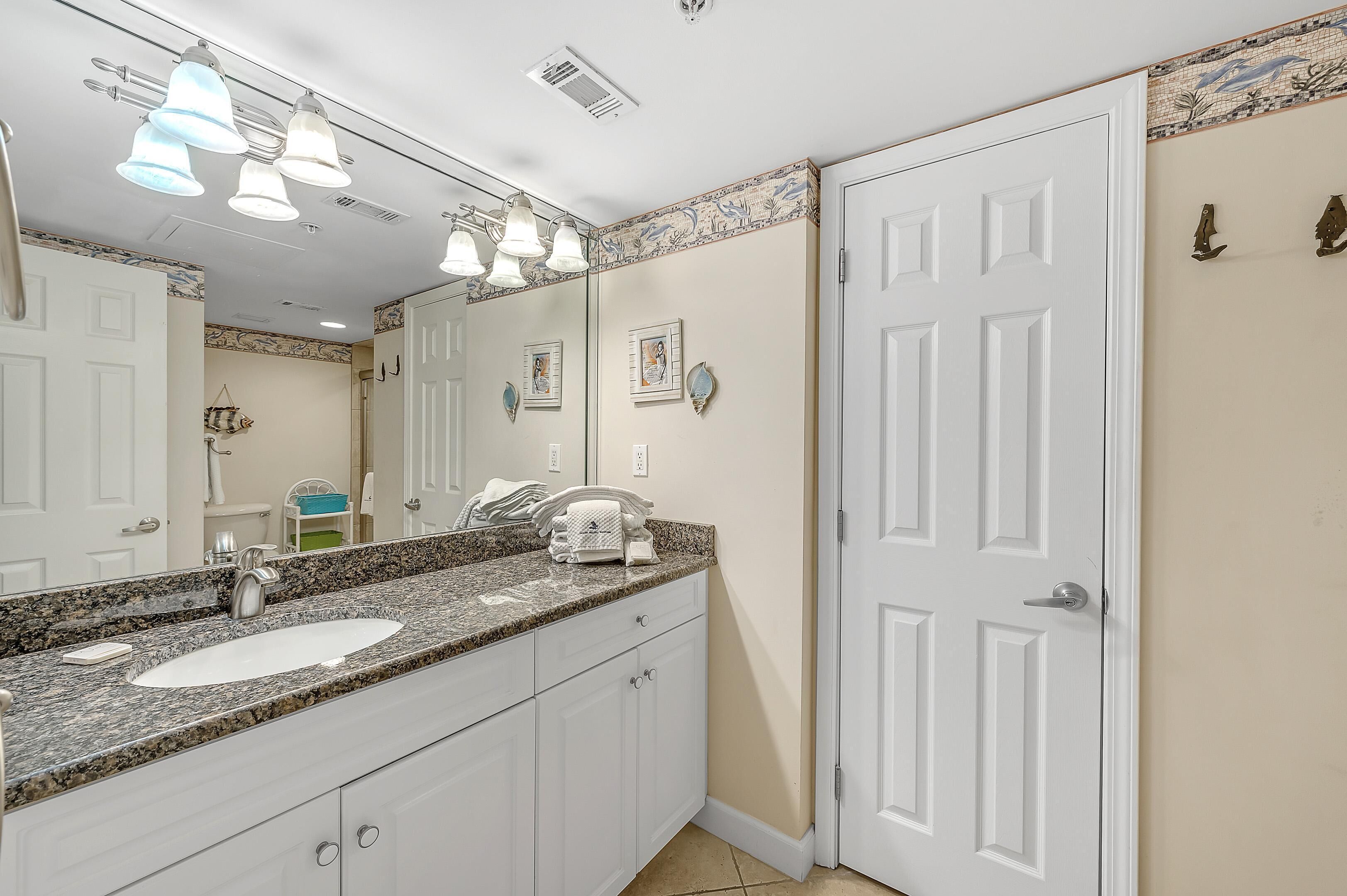 A bathroom with a white sink, granite countertop, and a large mirror. There are three overhead lights and a door with a decorative tile border.