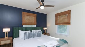 Une chambre avec une tête de lit recouverte d’un tissu vert, du linge de lit blanc et un ventilateur de plafond. On y trouve deux tables de chevet avec des lampes et une fenêtre avec un store en bambou.
