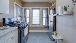 A compact kitchen with a stove, oven, and refrigerator. There are windows with curtains, a small table with a vase, and a shelf with items.