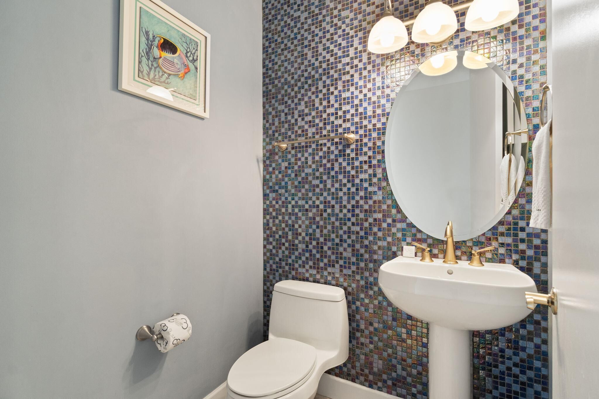 Une salle de bain avec un mur recouvert de mosaïques, des toilettes blanches, un lavabo avec des robinets dorés et un tableau encadré accroché au mur.