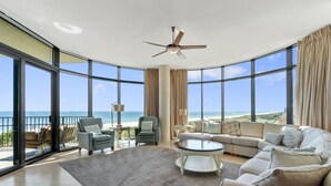 A spacious living room with a large window overlooking the beach, a ceiling fan, and a cozy seating area. - Capri 202 + Free Seasonal Beach Service (Pensacola)