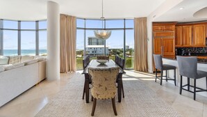 A modern dining area with a large window overlooking the ocean, a dining table with chairs, and a kitchen area with wooden cabinets. - Capri 202 + Free Seasonal Beach Service (Pensacola)