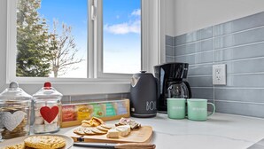 A kitchen counter with a variety of food items, including cookies, cheese, and a cutting board. There are two green mugs with the word "OCEANA" on them, a black coffee maker, and a glass jar with a heart decoration. - Whidbey Waterview Getaway: Nature, Deers, Pet & Family Friendly (Clinton)