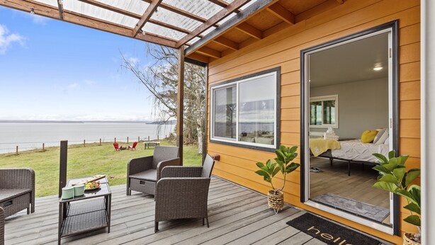 A wooden deck with wicker furniture, a view of the sea, and a bedroom with a bed and a window. - Whidbey Waterview Getaway: Nature, Deers, Pet & Family Friendly (Clinton)