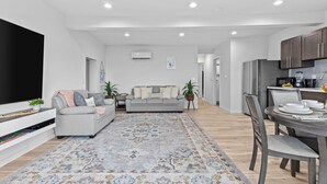 A modern living room with a grey sofa, a flat-screen TV, and a dining area with a table set for two. - Whidbey Waterview Getaway: Nature, Deers, Pet & Family Friendly (Clinton)