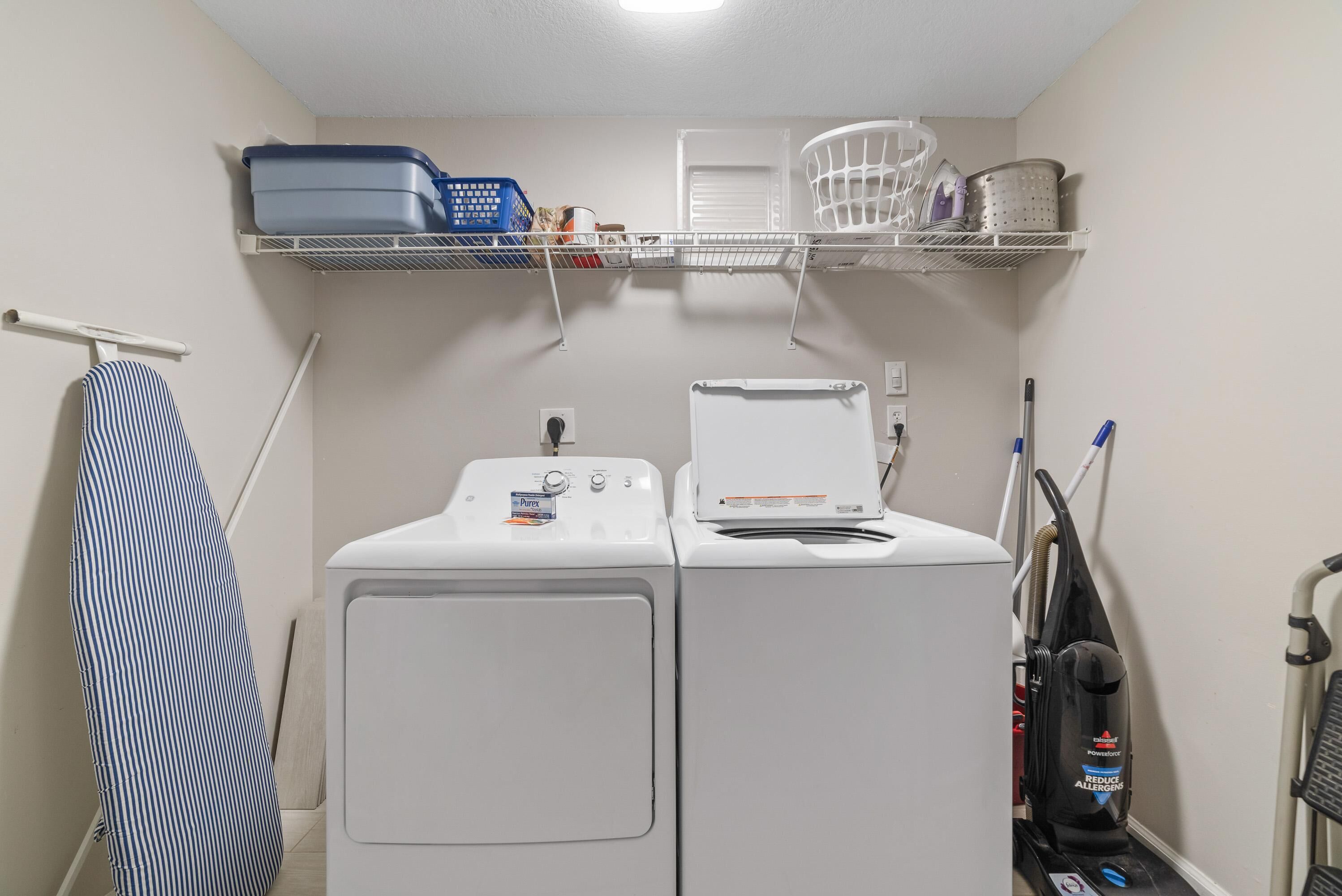 A laundry area with two washing machines, a drying rack, a vacuum cleaner, and storage shelves with baskets and containers.