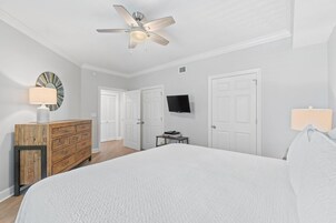 A bedroom with a large bed, a ceiling fan, a dresser, a TV, and two doors.