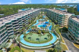 Um hotel com várias varandas, uma piscina central e uma área ajardinada com palmeiras.