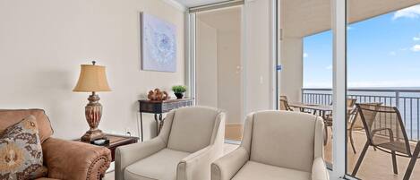 Quarto com sofá bege, duas poltronas bege, uma mesa com abajur e vista para a varanda com móveis de exterior.