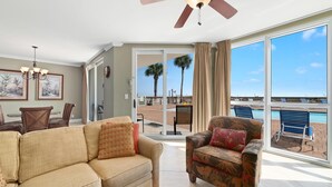 A living room with a sofa and armchair, a ceiling fan, and a view of the pool and palm trees outside. - Seaspray West 105- Gulf Front (Pensacola)