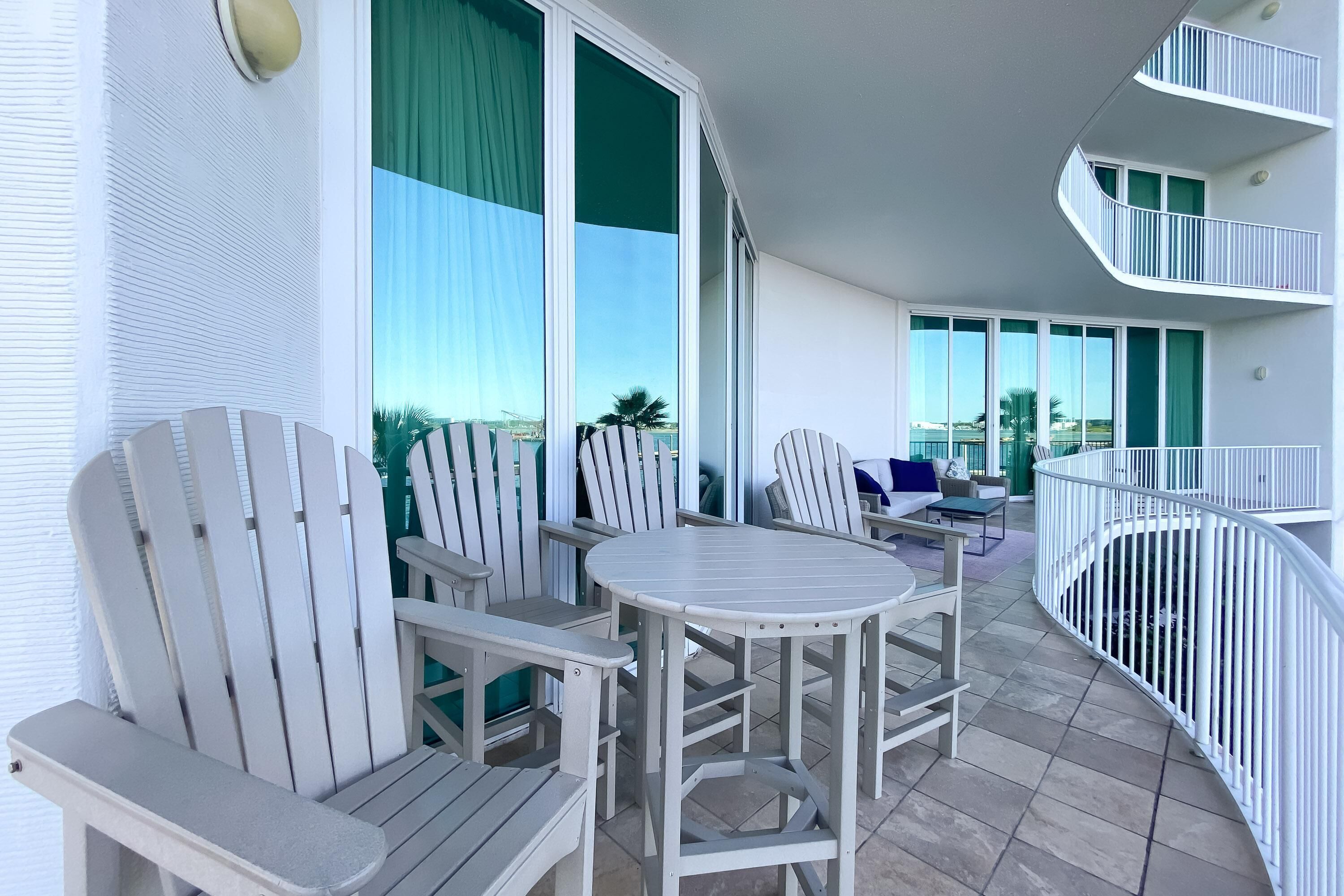 A balcony with white Adirondack chairs and a round table, large windows with curtains, and a spiral staircase.