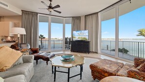 Een ruime woonkamer met een grote televisie, comfortabele zitmogelijkheden, een plafondventilator en een balkon met uitzicht op zee.