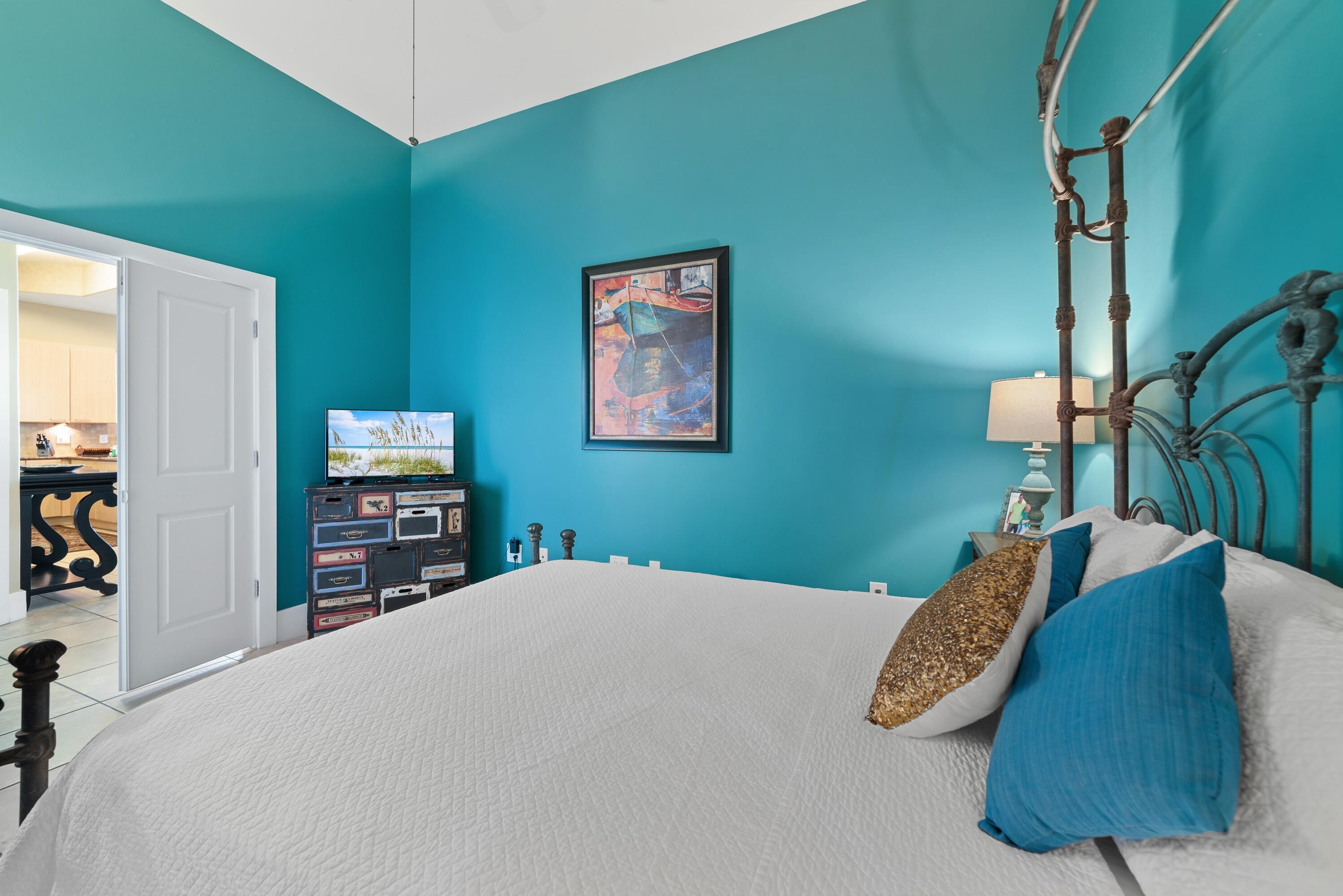 Un dormitorio con paredes verde azulado, una cama con ropa de cama blanca y almohadas azules, un cuadro abstracto enmarcado y un aparador con cajones.