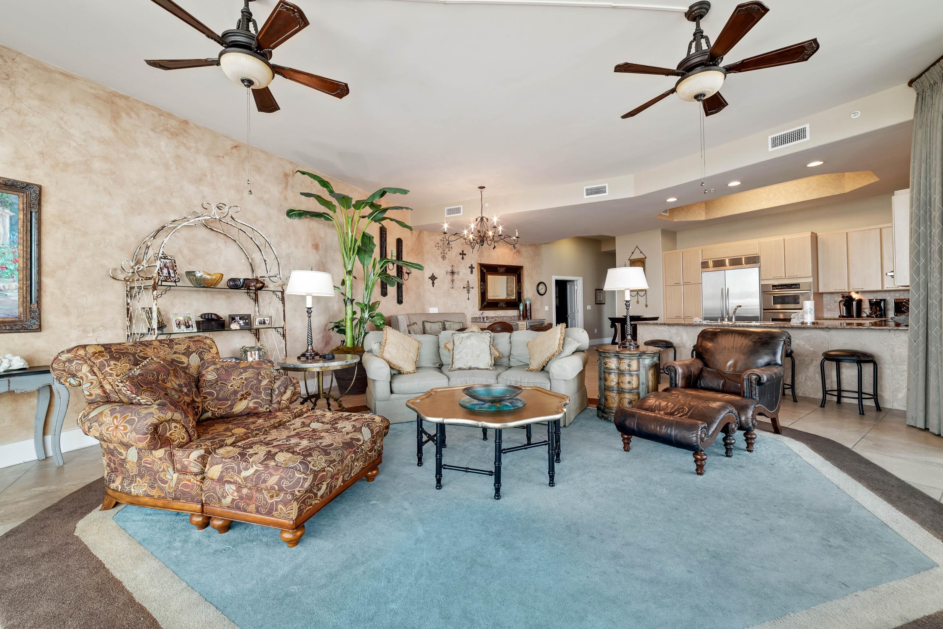 A spacious living room with a sofa, armchairs, and a coffee table. There are ceiling fans, a chandelier, and a kitchen in the background.