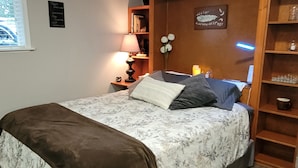 A bedroom with a bed, bedside tables, a lamp, a shelf with decorative items, and a wall with a sign.