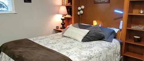 A bedroom with a bed, bedside tables, a lamp, a shelf with decorative items, and a wall with a sign.