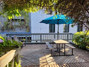 Une terrasse en bois avec une table et des chaises, un parasol bleu et, au fond, un bâtiment à la façade blanche.