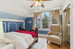 Une chambre à coucher avec un lit, une chaise, un ventilateur de plafond et une fenêtre avec des rideaux.