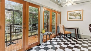 Ein Zimmer mit Schachbrettmuster-Boden, Deckenventilator, Tisch mit Stuhl und Blick ins Freie durch große Glasüren.