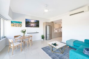 A modern living room with a dining area, a kitchen in the background, and a large window providing natural light.