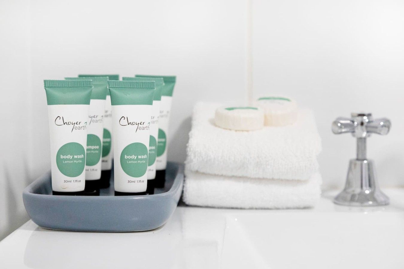 Une salle de bain avec un plateau gris sur lequel sont posés des flacons de gel douche, une pile de serviettes blanches et un robinet argenté.