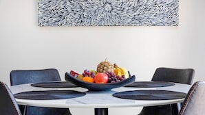 A round dining table with a black woven placemat, surrounded by four black leather chairs. A bowl of fruit is placed on the table.
