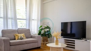 A living room with a grey sofa, a flat-screen TV, a small round table, and a potted plant. - Zen Nest: Cozy  ~ Lawn ~ Minimalist (Nightcliff)