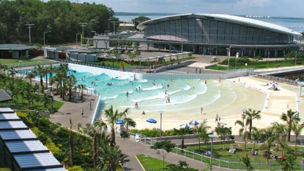 A large swimming pool with multiple lanes, a beach area with umbrellas, and a modern building with a curved roof.