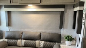 A modern RV interior with a sectional sofa, built-in cabinets, and a small table with a plant. - Beautiful NEW 2023 camper covered on concrete slab. Sit and enjoy the barges (Bolivar Peninsula)