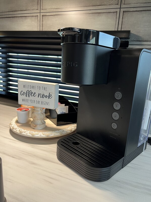 En Keurig-kaffemaskin står på en bänkskiva, och bredvid den finns en skylt med texten: ”Välkommen till vårt lilla kaffehörn, här börjar din dag.”