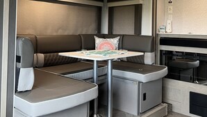 A compact RV interior with a dining area, seating, and a kitchenette. - Beautiful NEW 2023 camper covered on concrete slab. Sit and enjoy the barges (Bolivar Peninsula)