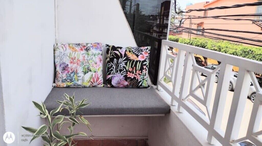 Uma varanda com um banco acolchoado, duas almofadas estampadas com flores e um vaso com planta.