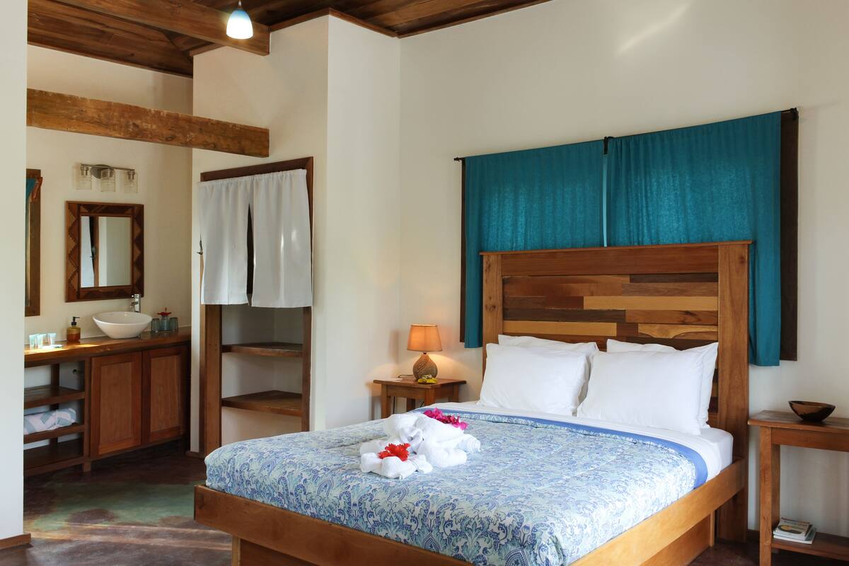 Une chambre avec un lit en bois, une table de chevet, une commode, un miroir, une salle de bain avec un lavabo et des placards, et une fenêtre avec des rideaux.